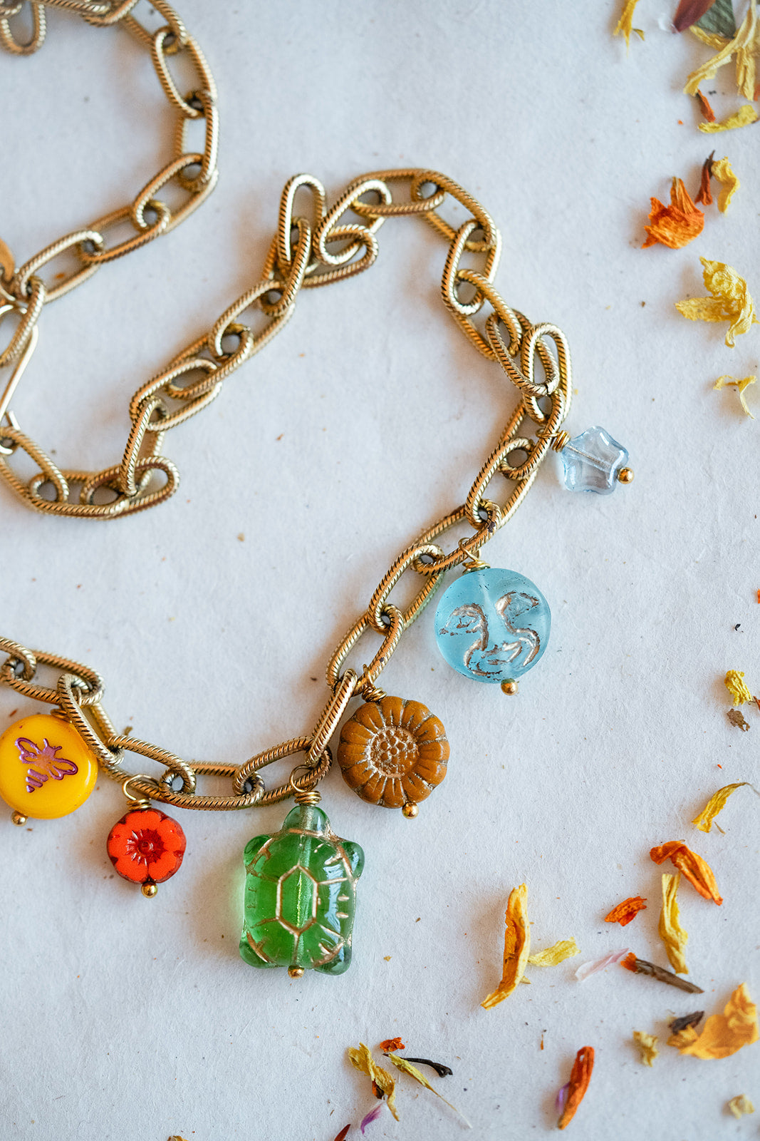 A gold-tone chain necklace with multiple colorful charms, including a green crystal pendant, a blue circular charm, a brown flower charm, and small orange and yellow floral accents, displayed on a white surface with scattered dried flower petals.