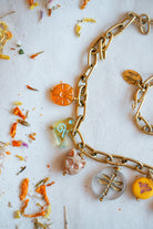A gold-toned chain necklace with multiple colorful charms, including a sun, a bird, a cat, a dragonfly, and a yellow flower, laid out on a white surface with scattered dried flowers.