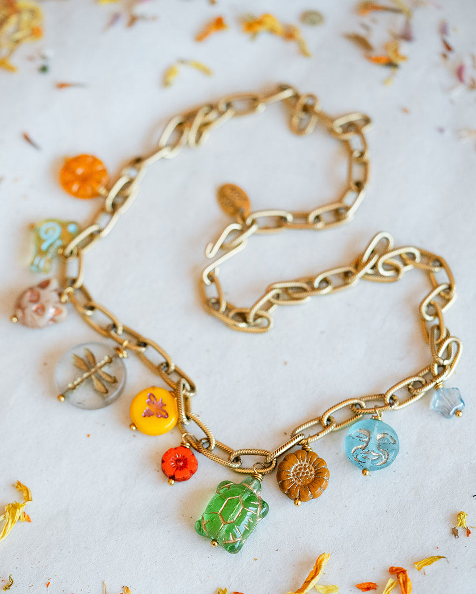 A gold chain necklace with multiple colorful enamel charms, including a green dragonfly, yellow sun, orange flower, and blue moon, arranged on a white surface with scattered dried flower petals.