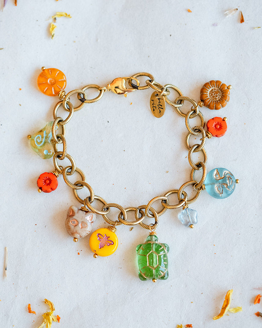 A gold-tone chain bracelet with colorful charm beads and pendants, laid flat on a white surface scattered with dried orange and yellow flower petals. The charms include orange, green, yellow, pink, and blue elements.