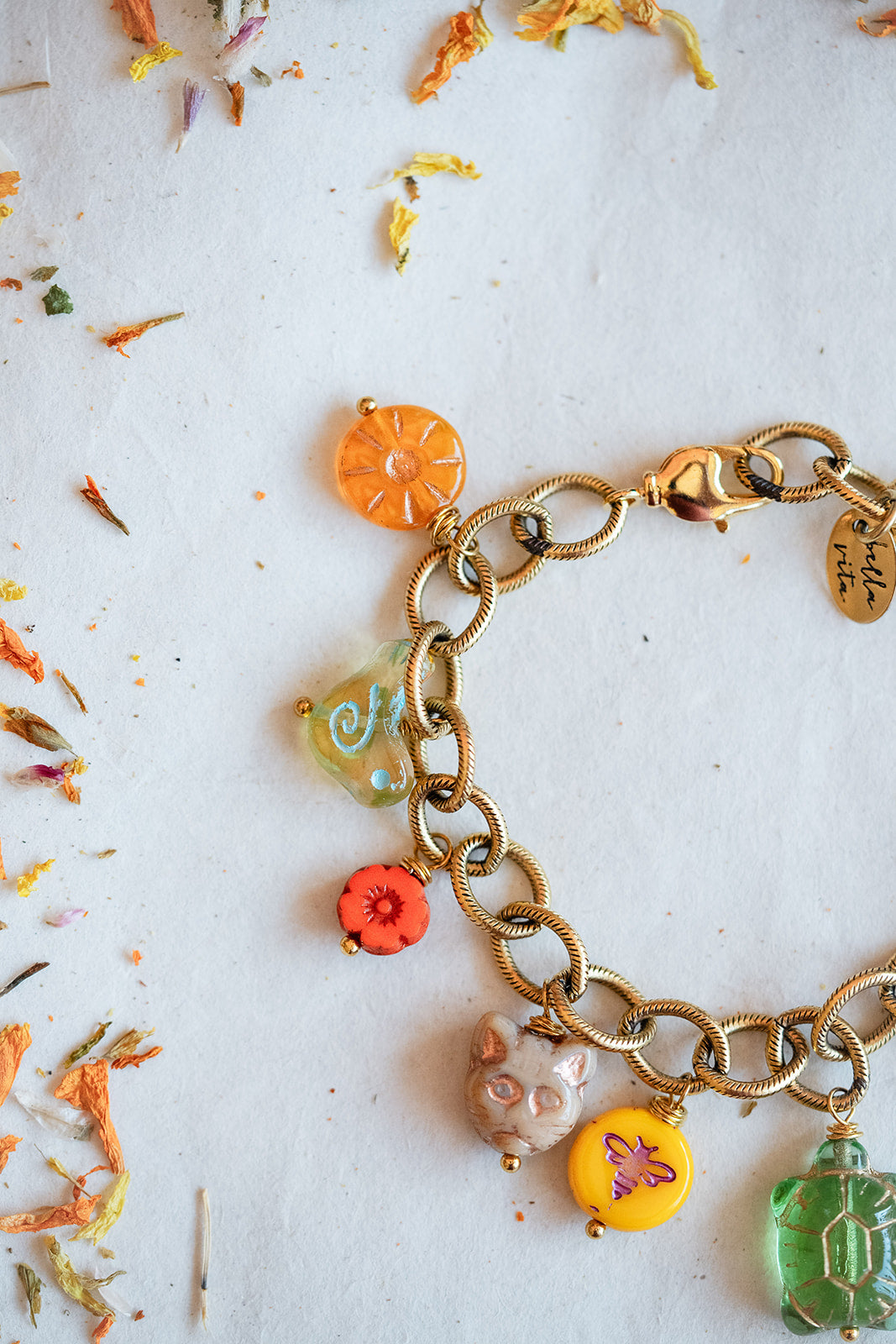 A gold-tone chain bracelet with colorful, whimsical charms including a cat, a butterfly, a flower, and a turtle, laid out on a white surface with scattered dried flower petals.