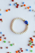 A beaded bracelet made of small off-white beads with a single blue button closure, arranged in a circle on a white background with scattered colorful beads around it.