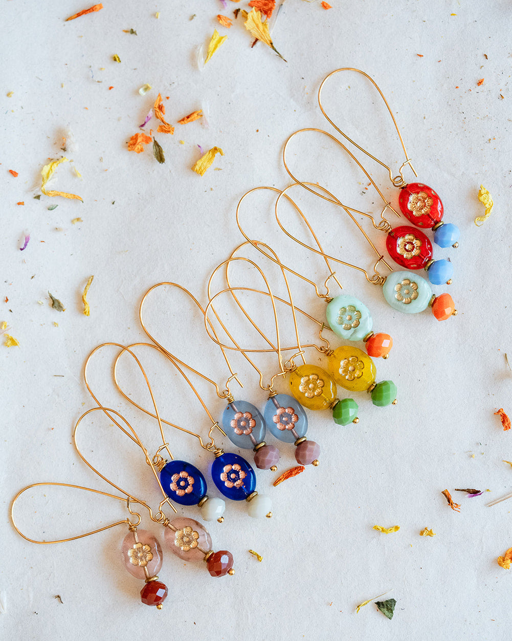 A set of gold-tone dangle earrings with colorful glass beads, arranged diagonally on a white surface scattered with dried flower petals. The earrings feature various bead colors including red, blue, yellow, green, pink, and light blue.