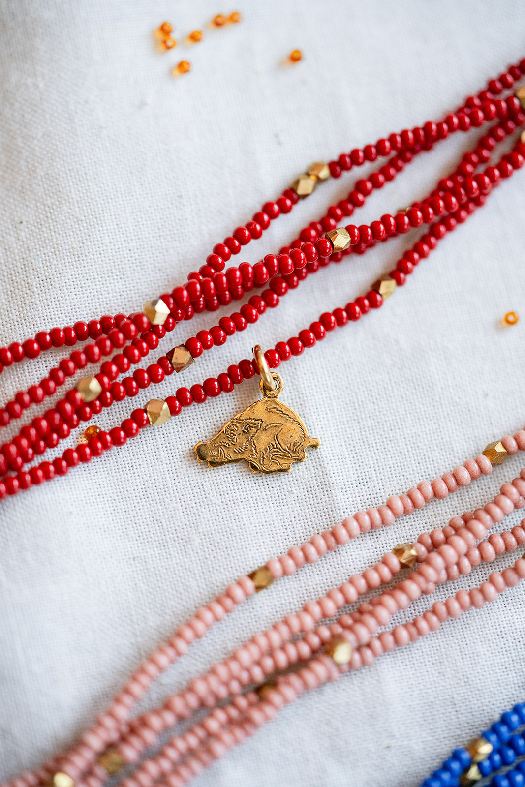 A close-up of multiple beaded necklaces laid out on a light fabric background. The most prominent necklace is made of bright red beads with small gold spacer beads and a small gold pendant.