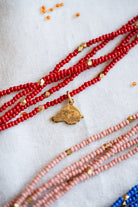 A close-up of multiple beaded necklaces laid out on a light fabric background. The most prominent necklace is made of bright red beads with small gold spacer beads and a small gold pendant.