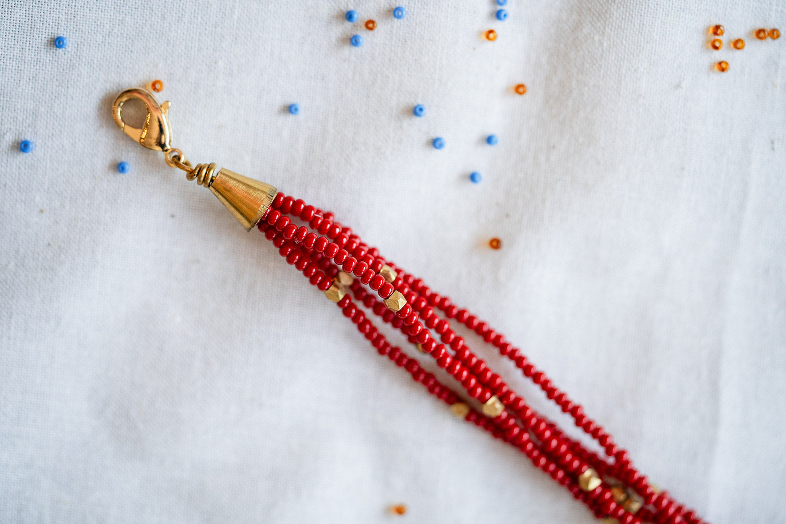 A close-up of a beaded tassel or strap end with multiple strands of red seed beads, a gold cone-shaped connector, and a gold lobster clasp, placed on a white fabric background with scattered small blue and orange beads.