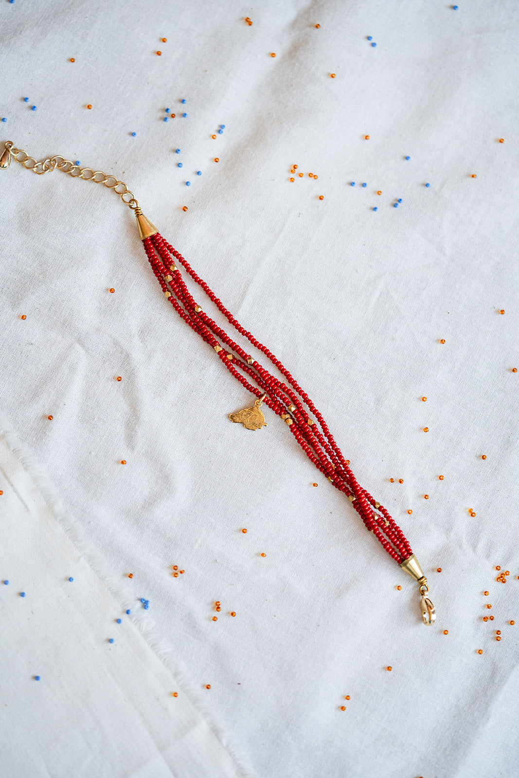 A beaded necklace with multiple strands of red beads, gold spacer beads, and a small gold pendant, laid flat on a white embroidered fabric background.