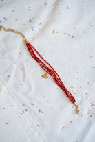 A beaded necklace with multiple strands of red beads, gold spacer beads, and a small gold pendant, laid flat on a white embroidered fabric background.