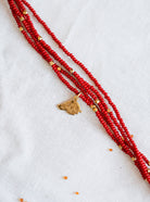 A multi-strand red beaded necklace with small gold spacer beads and a gold-toned pendant, laid diagonally on a white textured fabric background.