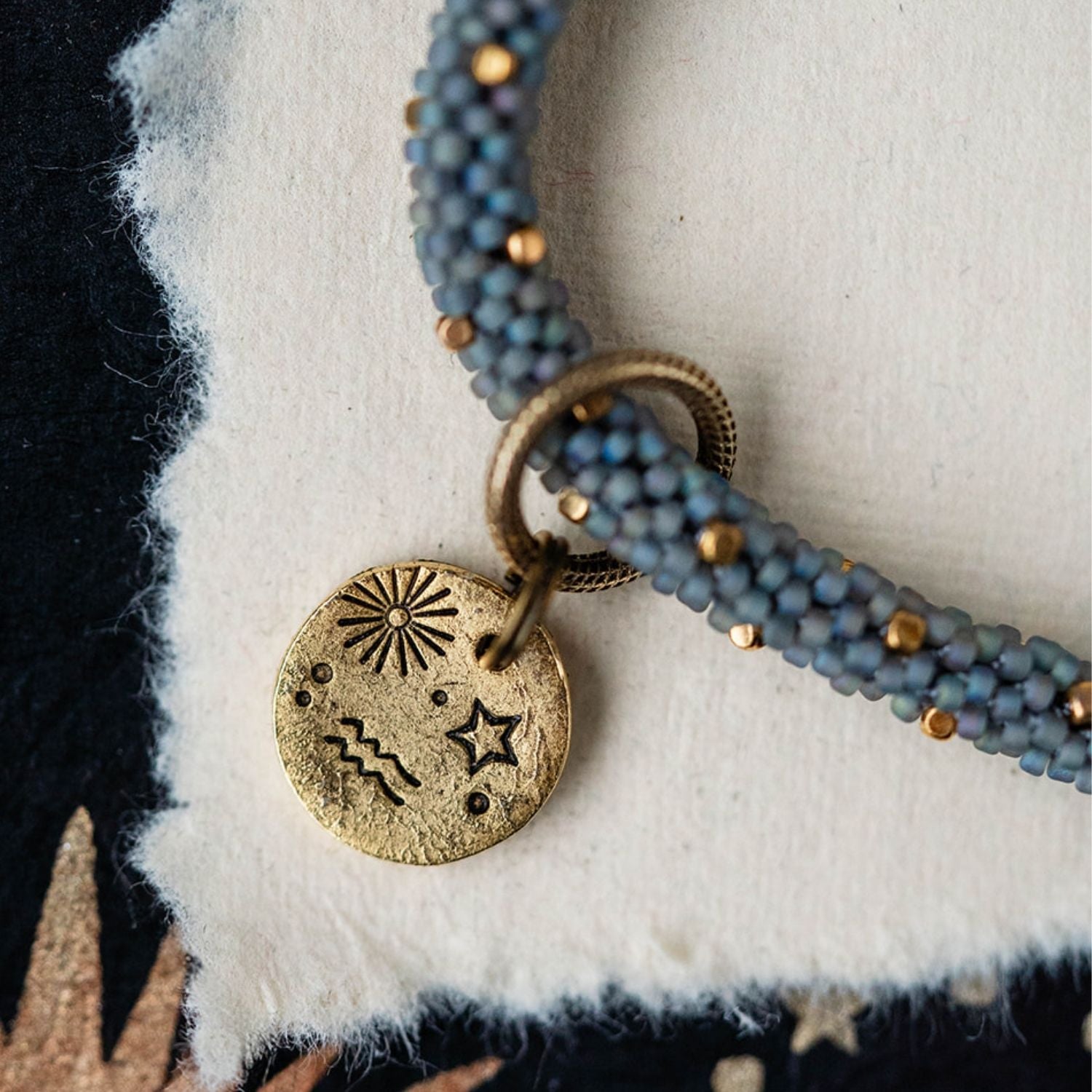 Gold Aquarius pendant attached to blue-grey beaded jewelry strands with gold accent beads detail view