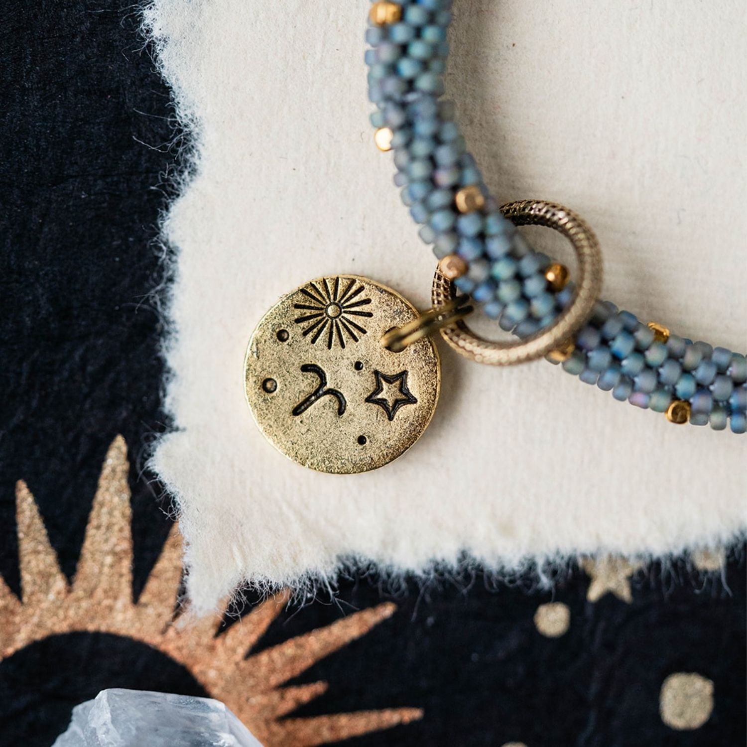 Gold Aries charm with blue-grey beaded strands showing sun and ram horn celestial engravings