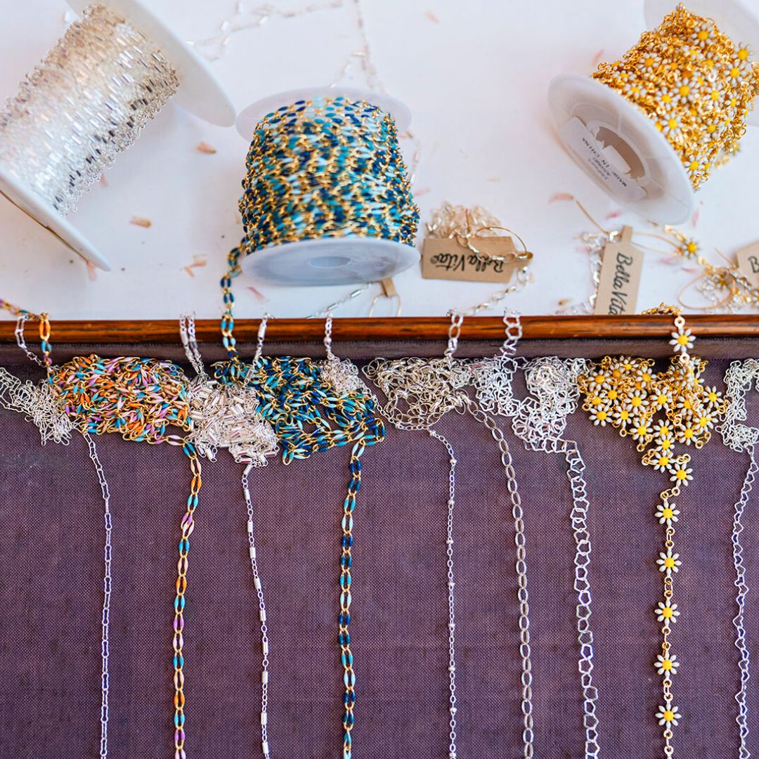 Bella Vita's permanent jewelry chain collection featuring enamel-coated chains in turquoise and rainbow colors, sterling silver link chains, and gold daisy chain designs on display with inventory spools