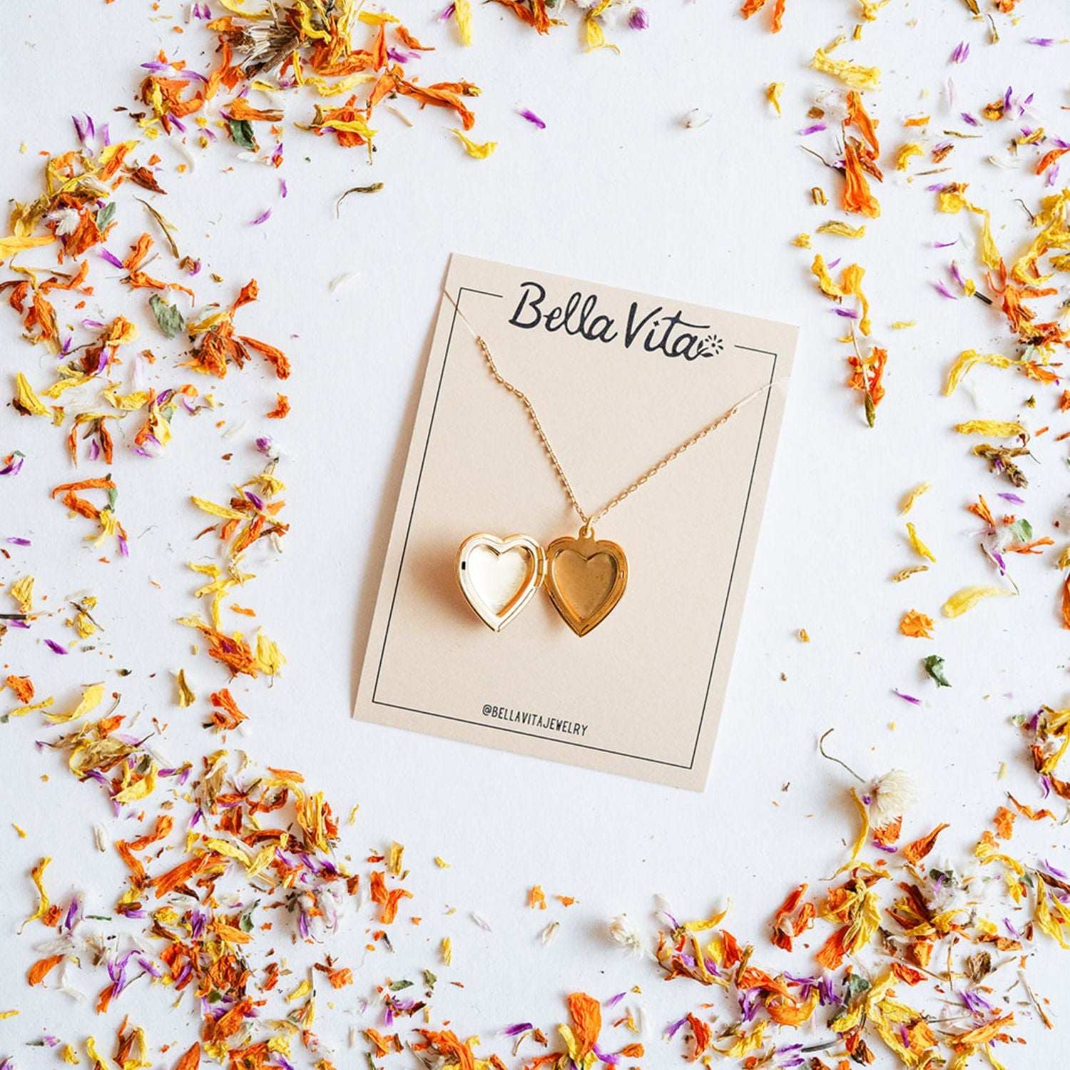 Handcrafted gold heart locket necklace from Bella Vita showing the open locket design on a branded card, surrounded by vibrant dried flower petals, perfect for keeping memories close.