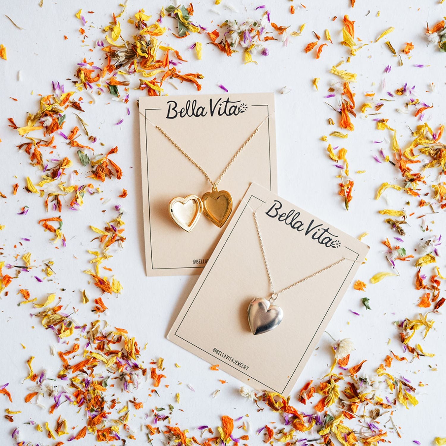 Bella Vita's handcrafted heart necklace collection featuring an open gold locket and solid silver heart pendant displayed on branded cards, surrounded by colorful dried flower petals, all made by hand in Little Rock.