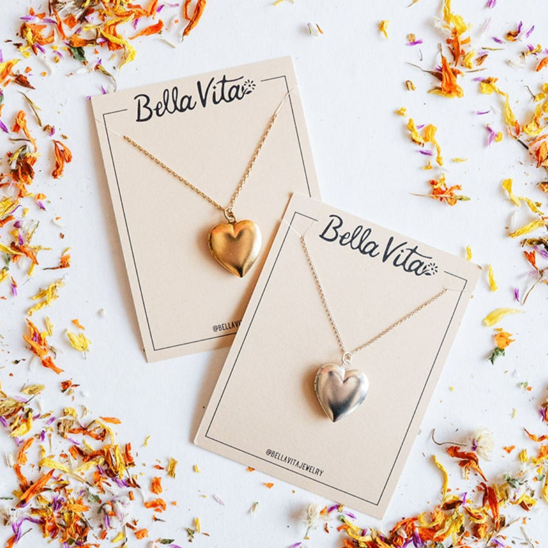 Bella Vita's handcrafted heart locket necklaces in gold and silver finishes displayed on branded cards surrounded by colorful dried flower petals, each made by hand in our Little Rock studio.