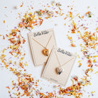 Bella Vita's handcrafted heart locket necklaces in gold and silver finishes displayed on branded cards surrounded by colorful dried flower petals, each made by hand in our Little Rock studio.