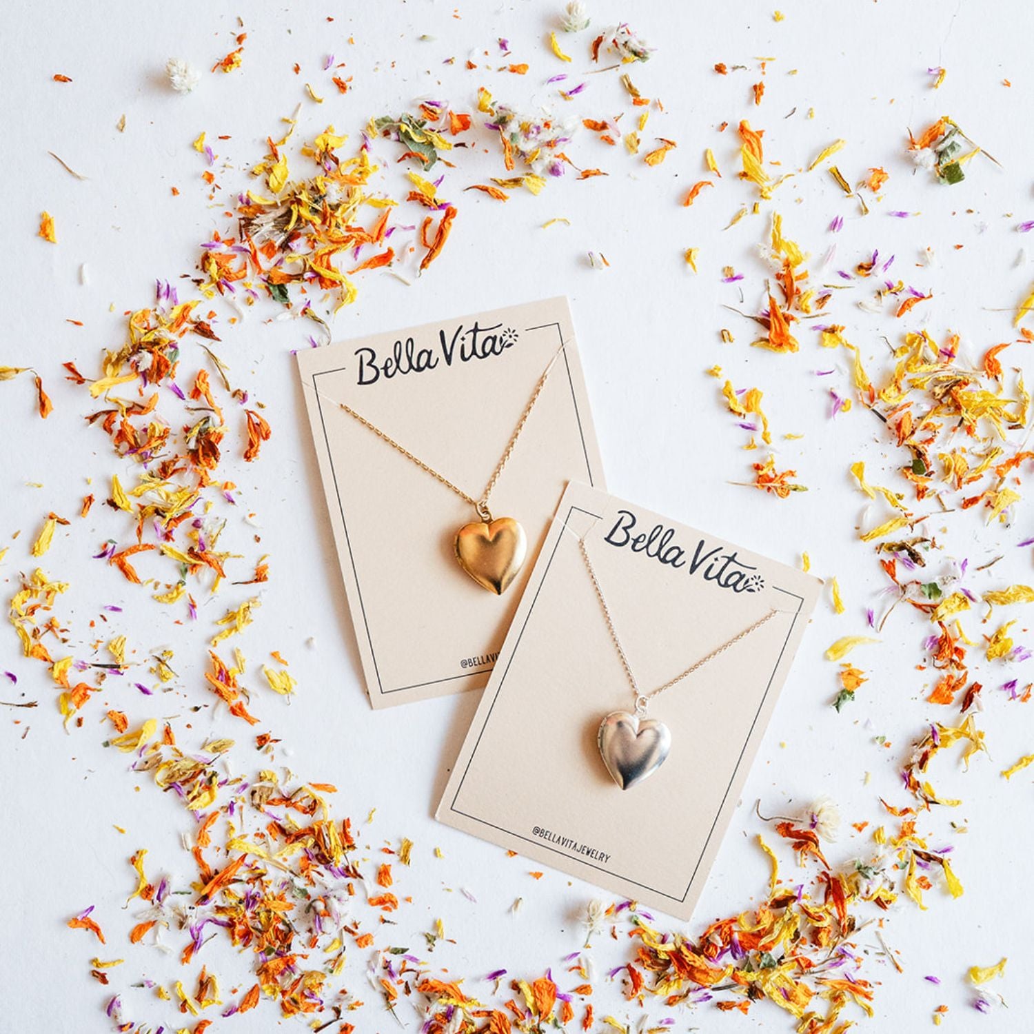 Bella Vita's handcrafted heart locket necklaces in gold and silver finishes displayed on branded cards surrounded by colorful dried flower petals, each made by hand in our Little Rock studio.