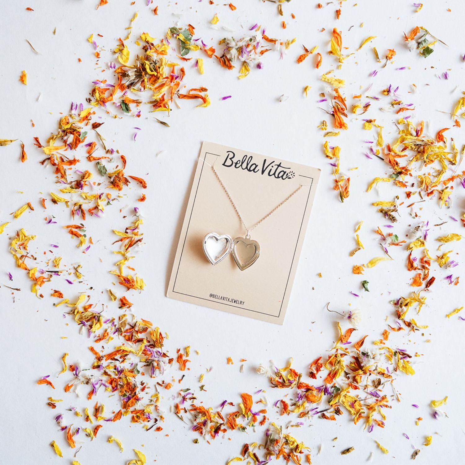 Bella Vita's silver heart locket necklace shown with open locket design on a branded card, surrounded by vibrant dried petals, handcrafted to hold personal mementos and photos.