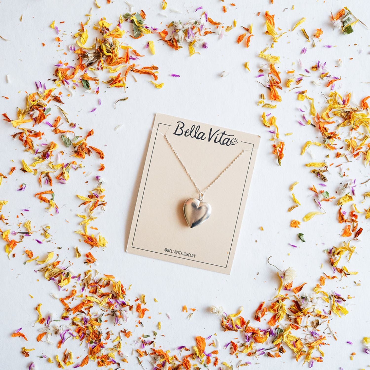 Sterling silver heart locket necklace handmade by Bella Vita artisans, presented on a branded card surrounded by a halo of colorful dried flower petals, creating a meaningful gift.
