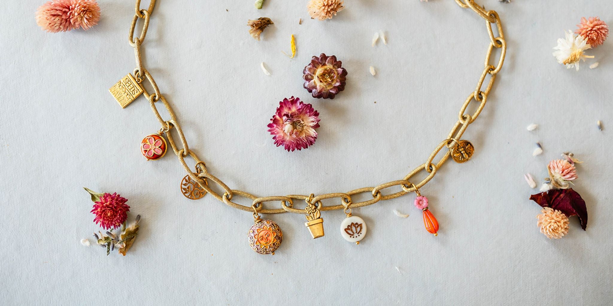 Handcrafted gold chain charm bracelet from Bella Vita's Garden Collection featuring lotus symbol, flower charms, and golden pot charm, styled with dried strawflowers in pink and burgundy