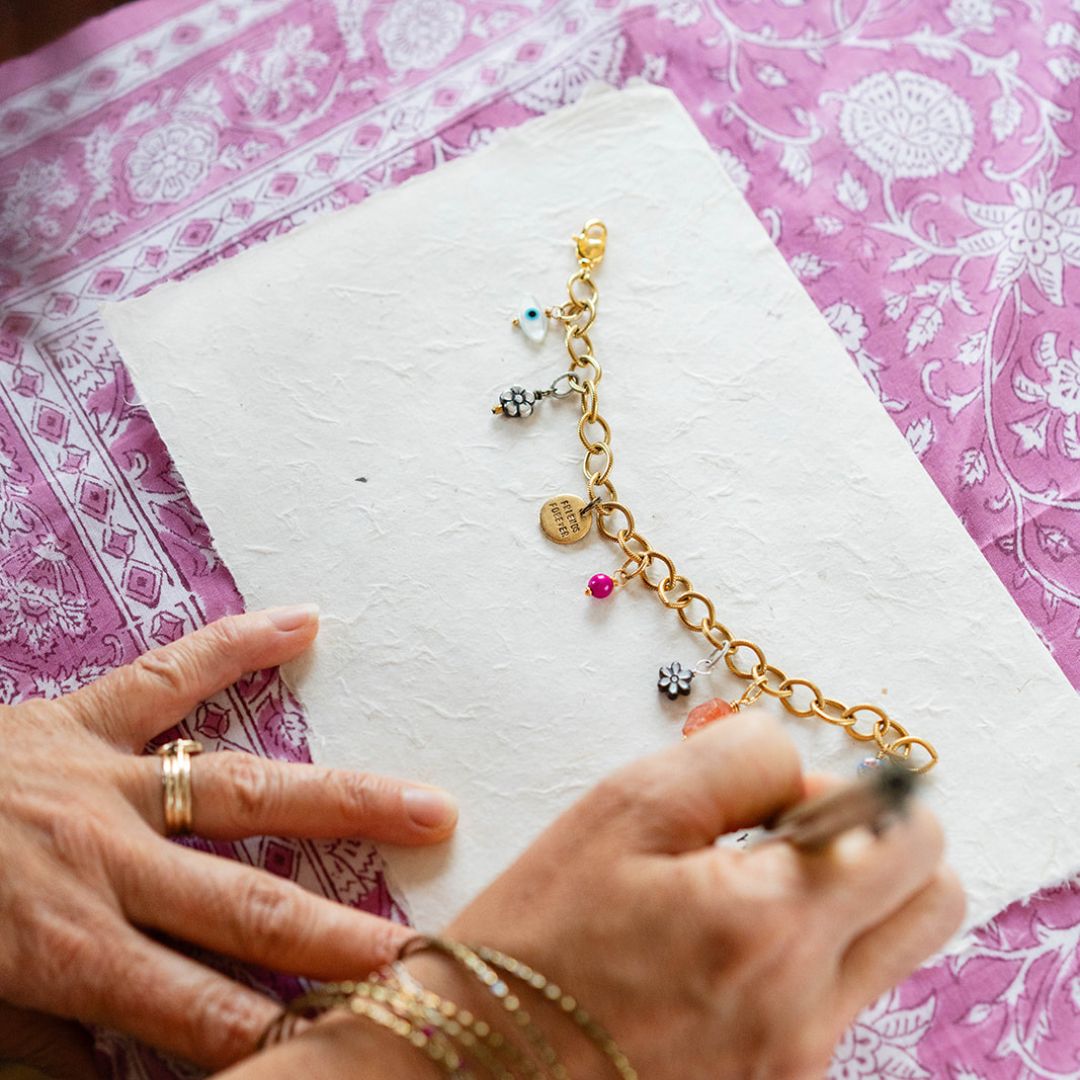 Artisan creating personalized charm bracelet with gold chain and mixed metal flower charms on pink floral fabric