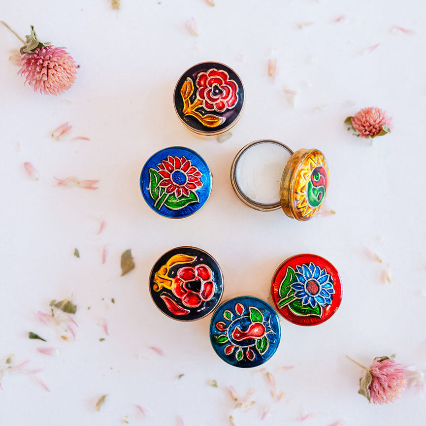 Flatlay collection of Song of India decorative brass solid perfume containers showing varied artistic designs with floral motifs in black, blue, yellow and red, styled with dried flowers.
