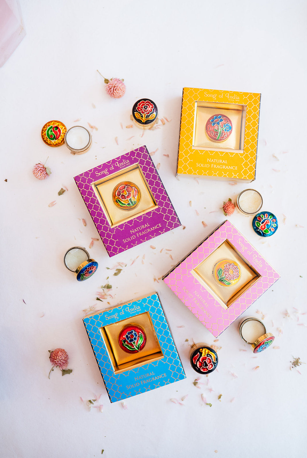 Flatlay arrangement of Song of India complete solid perfume collection showing colorful boxes in pink, blue, yellow and purple with opened brass containers and floral accents