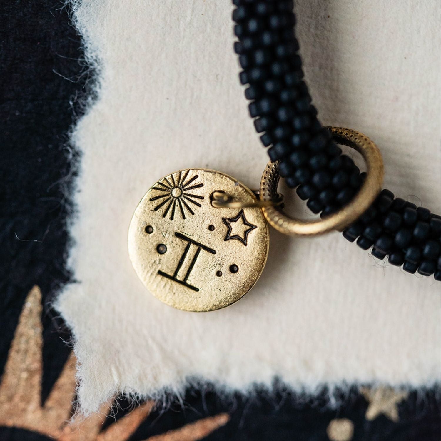 Close-up gold Gemini zodiac charm with twins symbol attached to black beaded jewelry detail