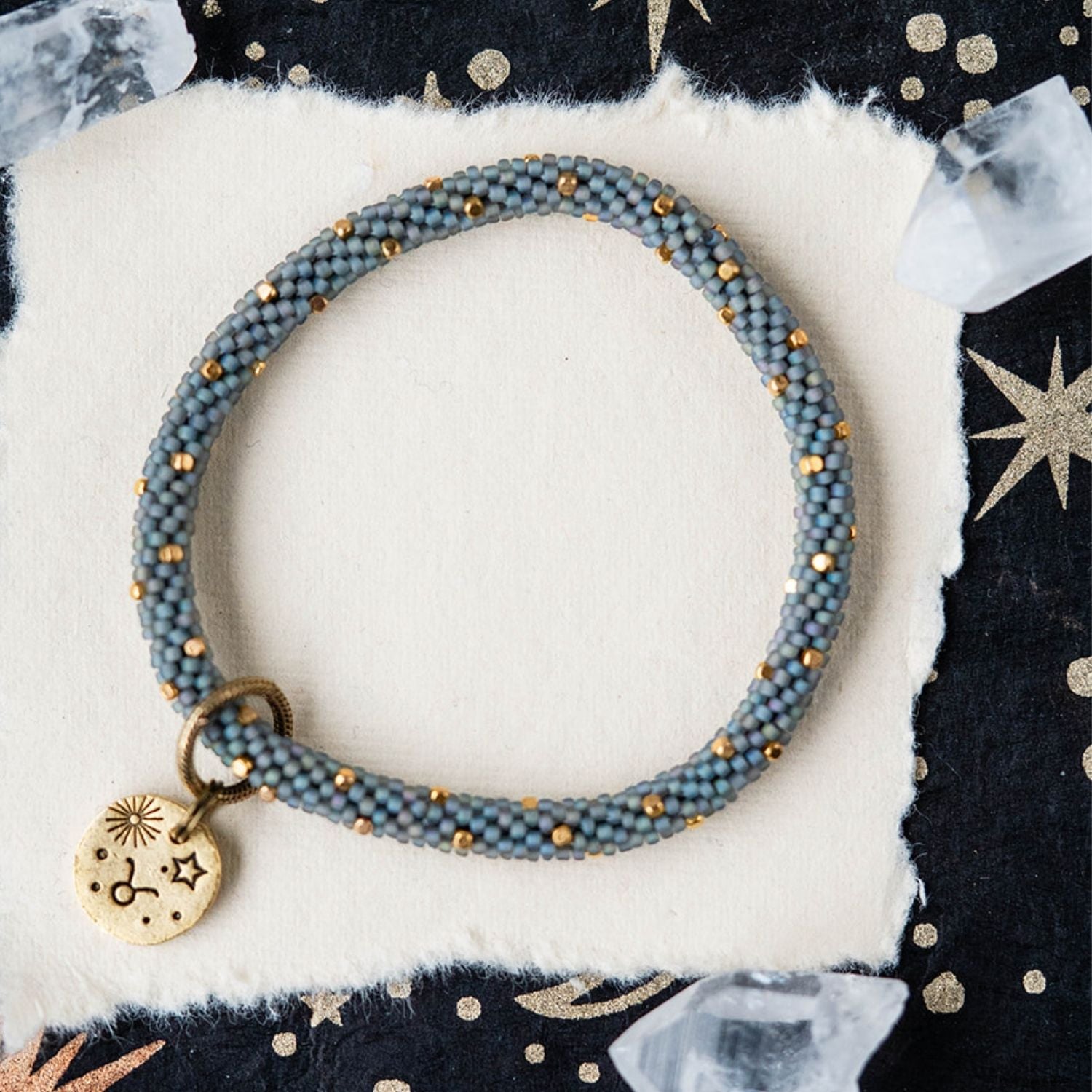 Blue-grey beaded bracelet with gold spacers and Taurus zodiac charm on starry background