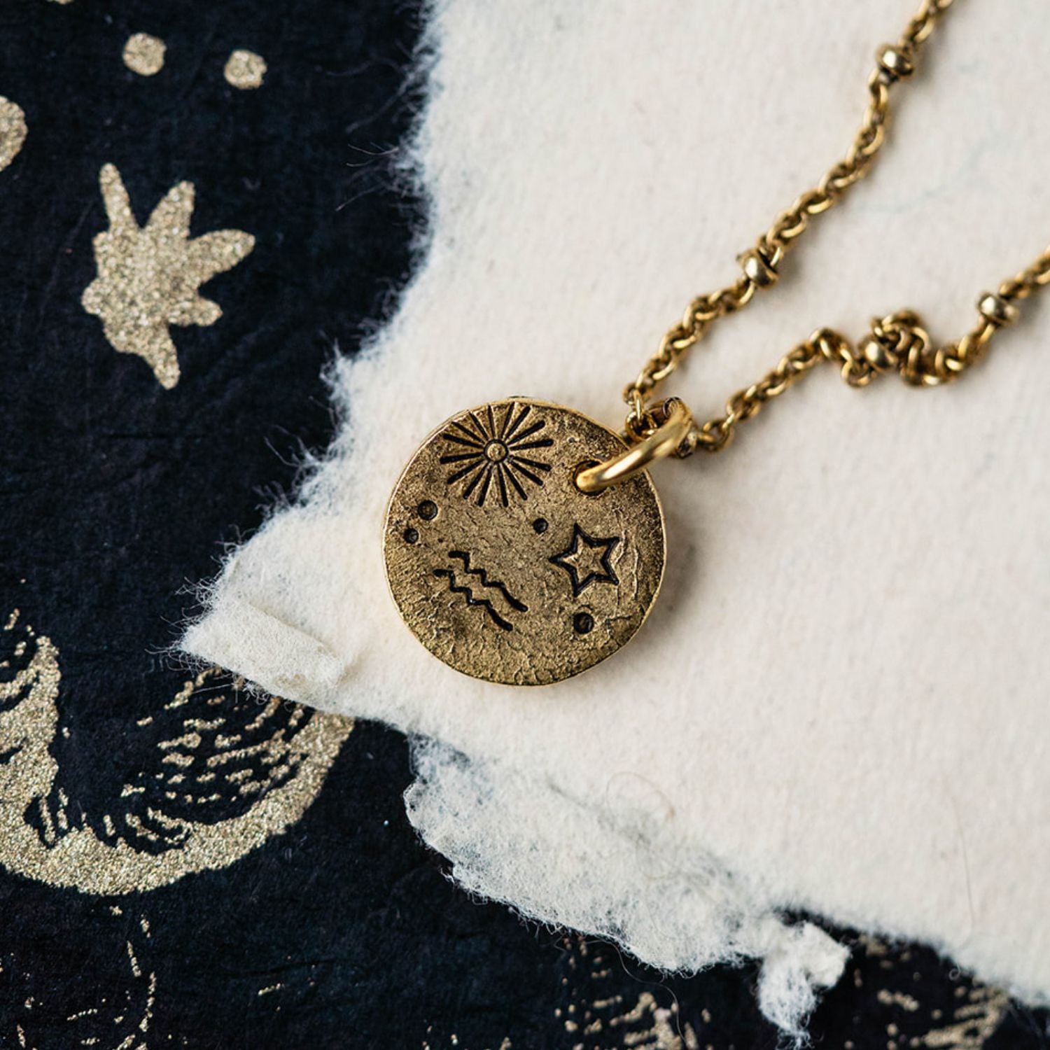 Gold Aquarius zodiac pendant necklace with celestial symbols on chain against starry fabric background