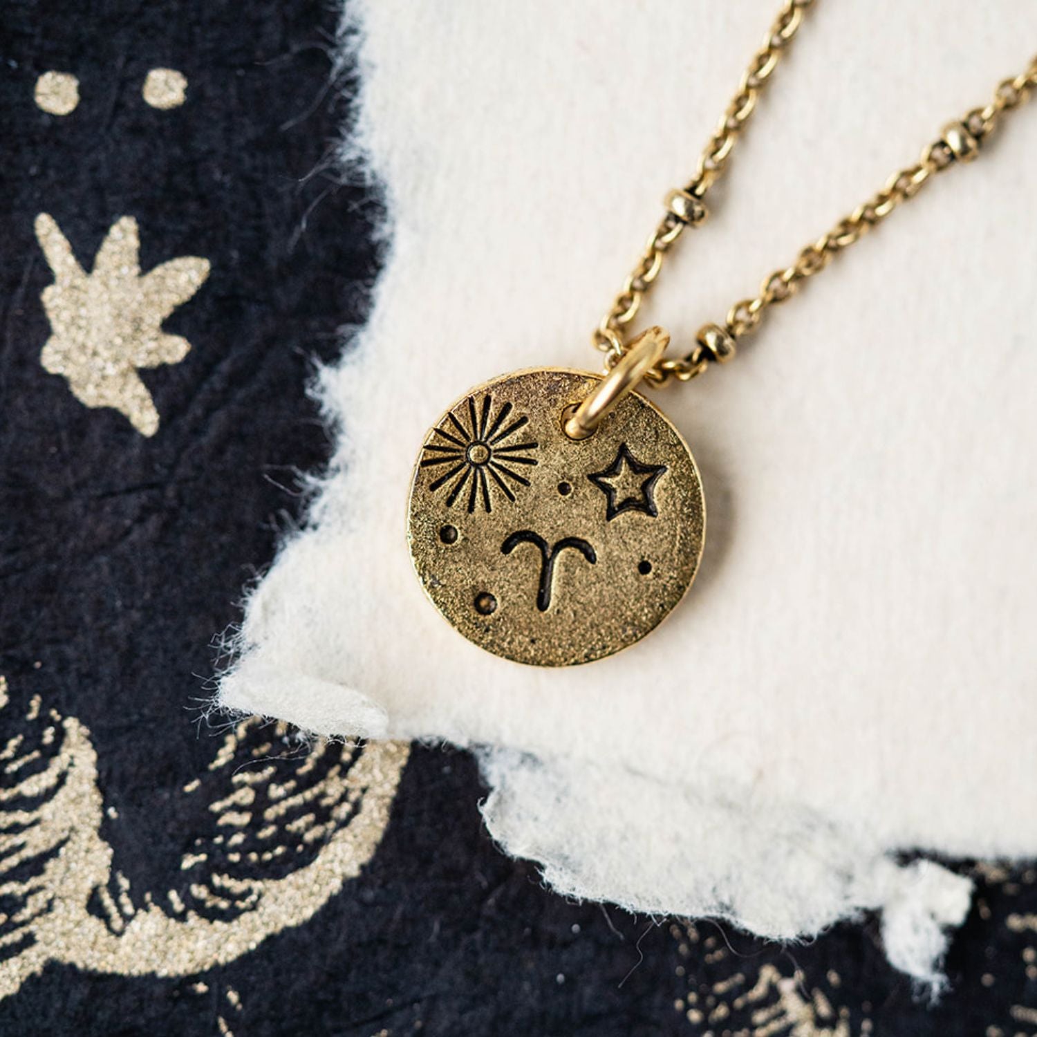 Gold Aries zodiac pendant necklace with ram horn and sun symbols on celestial fabric background